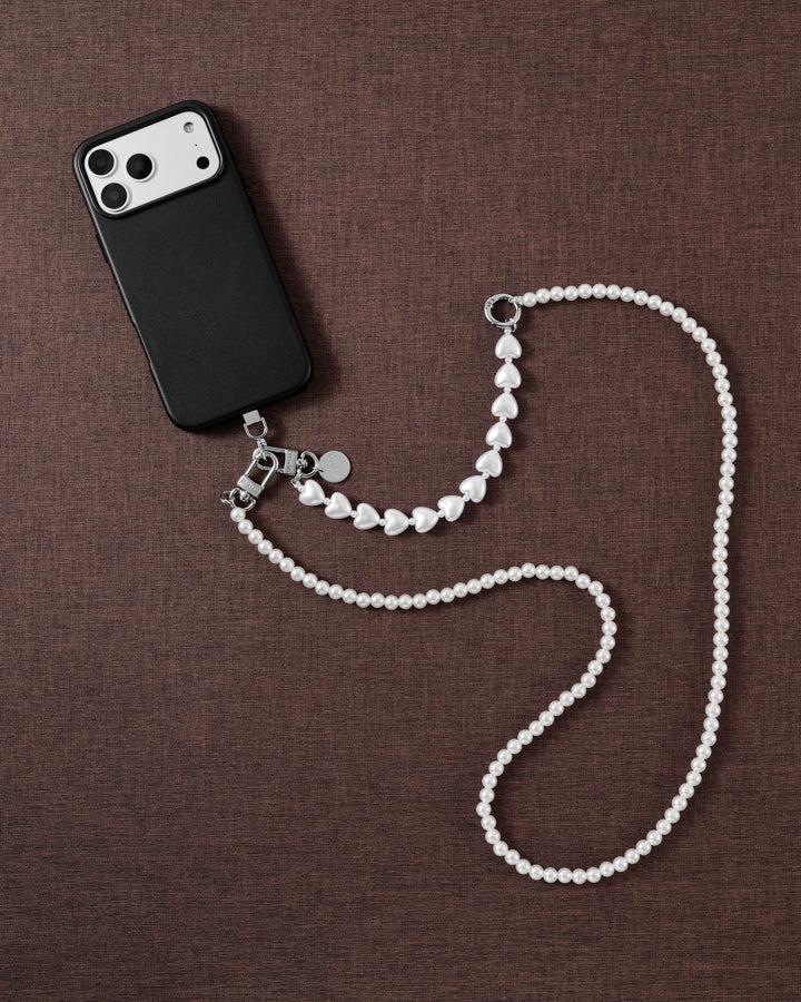 Styled flat-lay on brown fabric: a black phone with the white pearl-and-heart strap draped crossbody-length, silver-tone clasp and disc.