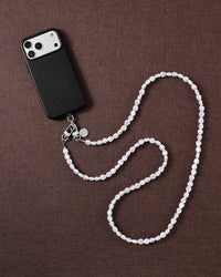 Flat-lay of a black phone and the irregular white faux-pearl cross-body strap curving across a warm brown fabric backdrop.