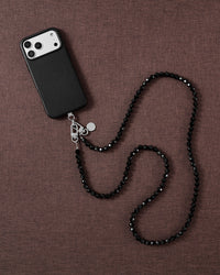 Styled flat-lay on mauve linen: a black-cased phone with a faceted jet-black beaded strap curving across the fabric.