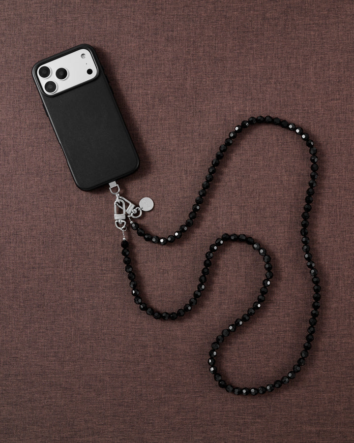 Styled flat-lay on mauve linen: a black-cased phone with a faceted jet-black beaded strap curving across the fabric.