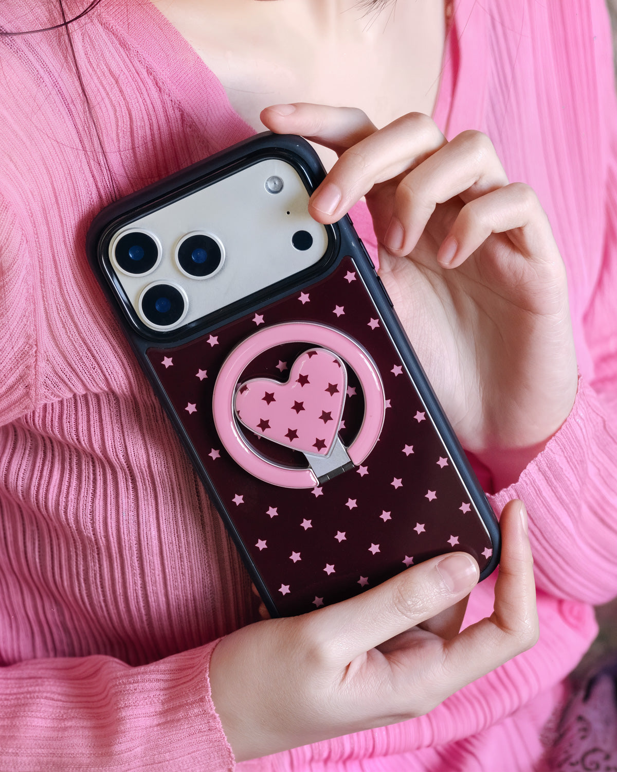 Phone propped between fingers with the pink heart ring kickstand visible on the burgundy case back