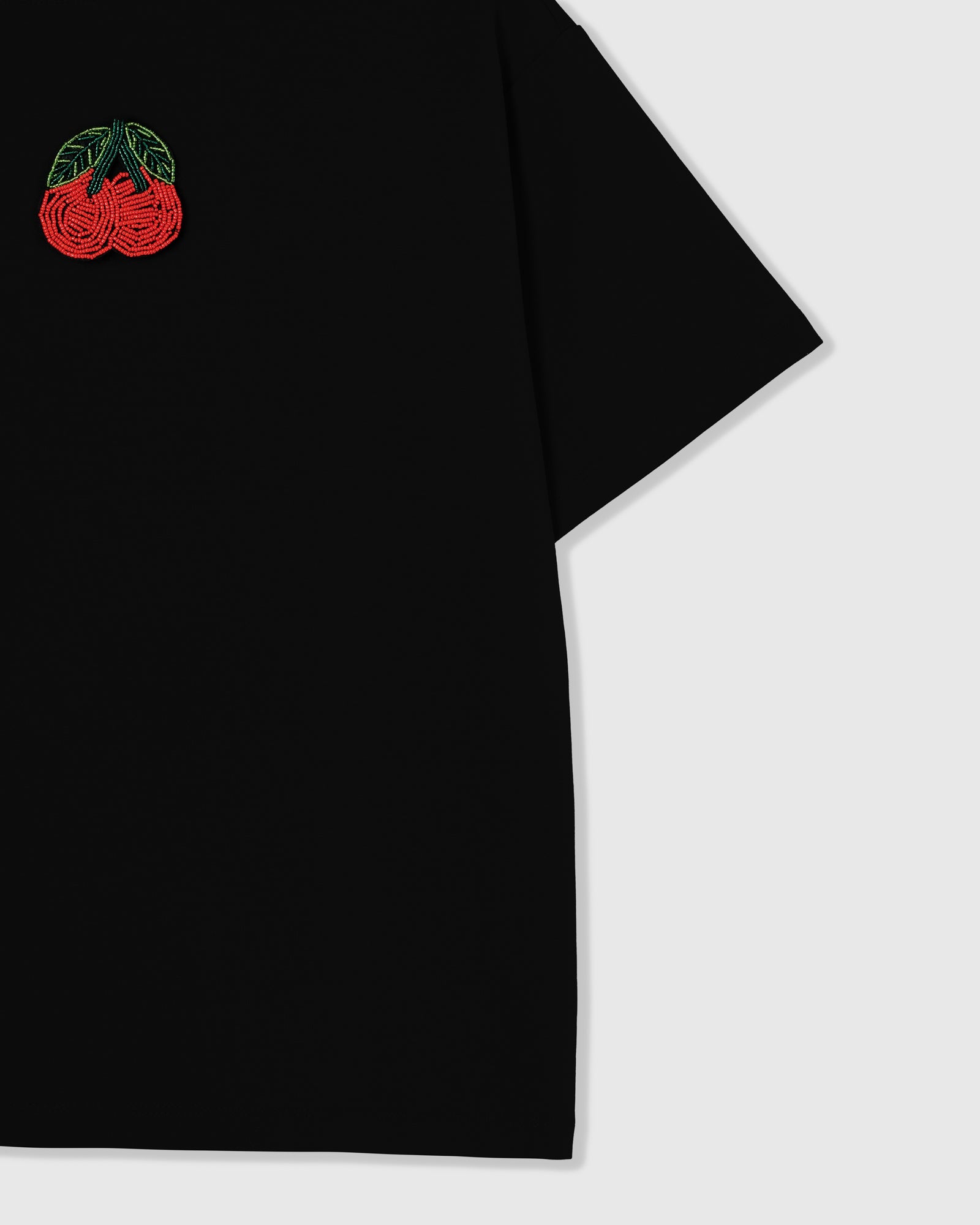 Close-up of the hand-beaded cherry motif sitting raised against the black cotton tee, with the sleeve in frame