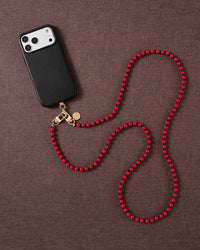 Styled flat-lay of a black phone clipped to the scarlet beaded cross-body strap, coiled in loops on a warm brown textured fabric.