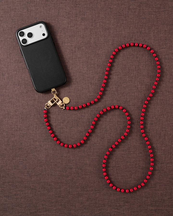 Styled flat-lay of a black phone clipped to the scarlet beaded cross-body strap, coiled in loops on a warm brown textured fabric.