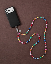 Styled flat-lay of a phone and the rainbow heart-bead cross-body strap coiled on warm brown fabric, showing its full over-the-shoulder length.