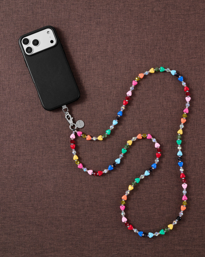 Styled flat-lay of a phone and the rainbow heart-bead cross-body strap coiled on warm brown fabric, showing its full over-the-shoulder length.