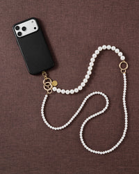 Styled flat-lay on brown fabric: black phone case and the full two-size faux-pearl cross-body strap with gold-tone ring and clasp