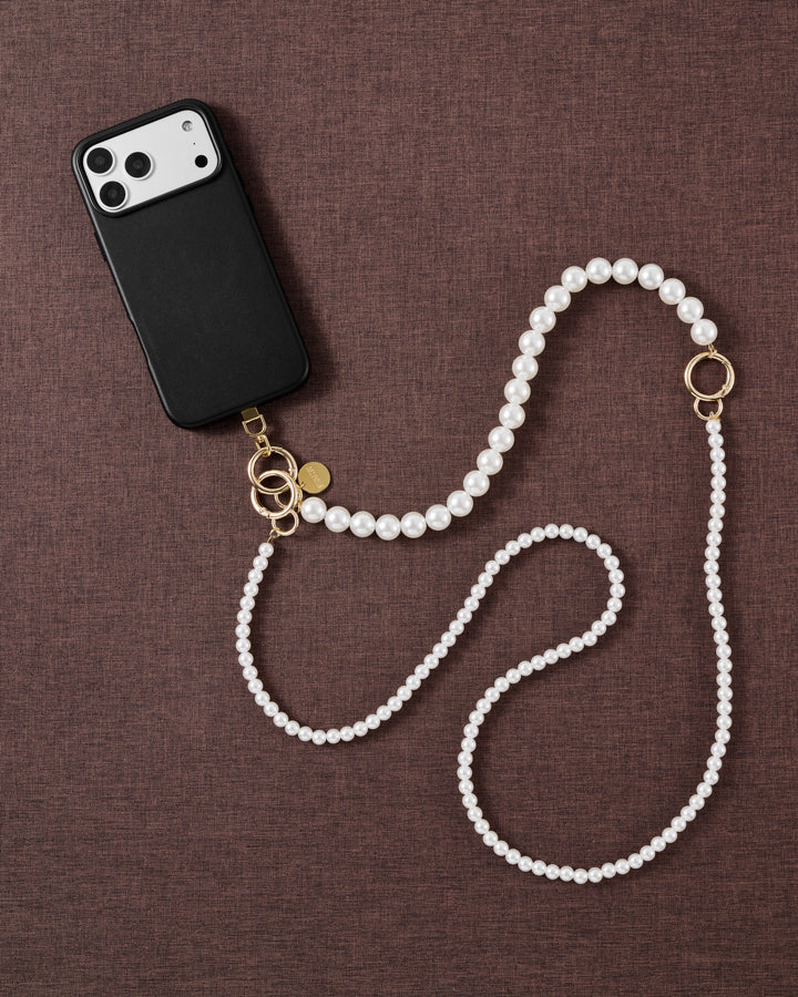 Styled flat-lay on brown fabric: black phone case and the full two-size faux-pearl cross-body strap with gold-tone ring and clasp