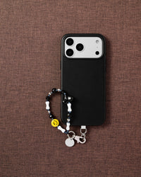 Black smiley heart phone strap flat-lay on dark brown fabric by STICKYZOO