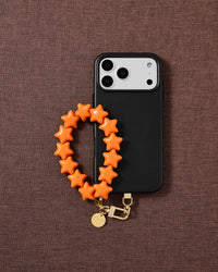 Tangerine Star phone strap and black phone case styled on warm brown fabric — daily wrist carry.