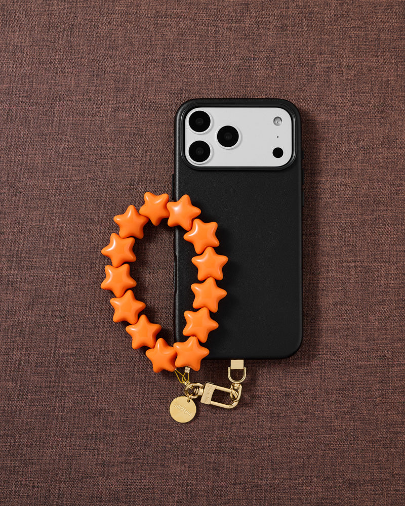 Tangerine Star phone strap and black phone case styled on warm brown fabric — daily wrist carry.