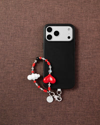 Phone with red heart and cloud charm strap on dark brown textured fabric lifestyle flat-lay
