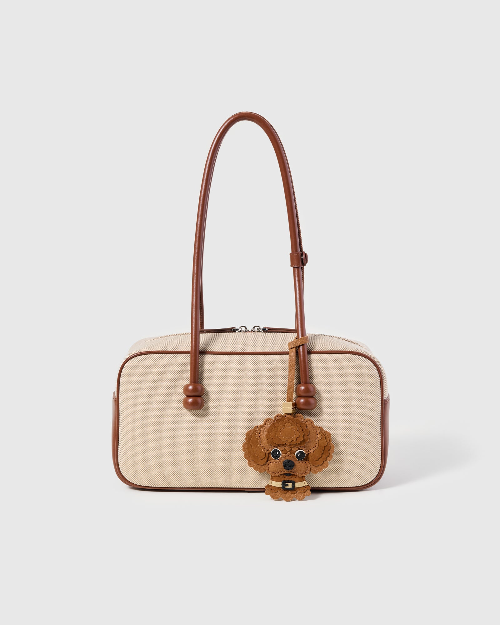 {{A detail people notice.}} The Poodle Charm styled on a cream canvas-and-tan top-handle bag, front — the exclusive drop that changes the way a bag feels.