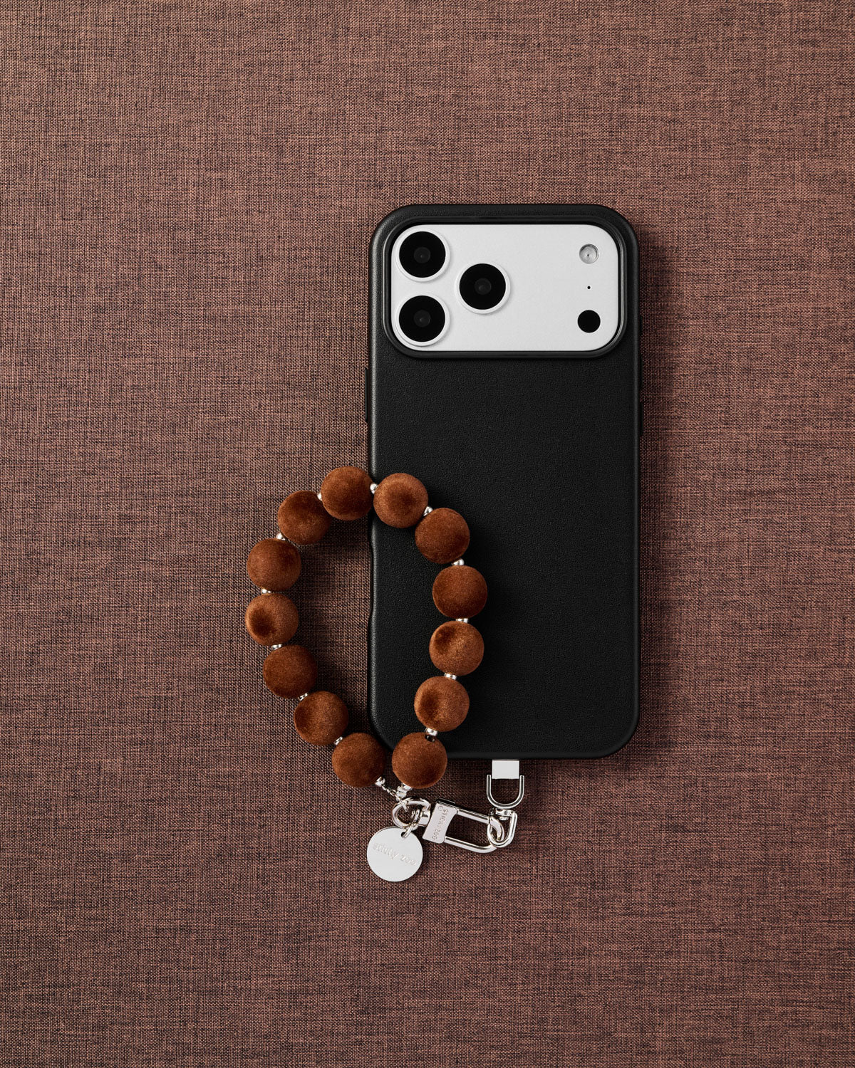 {{Designed in Amsterdam}} Phone with camel velvet pom bead strap on dark brown fabric lifestyle flat-lay