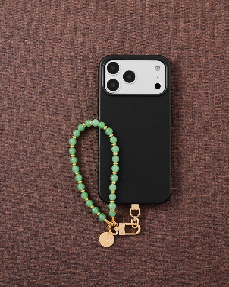 Phone with sage green orbit bead strap on dark brown fabric lifestyle flat-lay