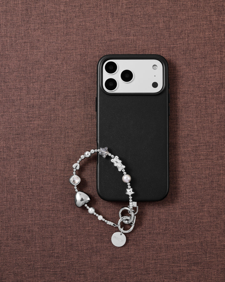 Silver heart-and-star beaded phone wrist strap draped on a black case over mauve linen, with a clear faceted bead, grey faceted star, grey pearl-effect bead and disc