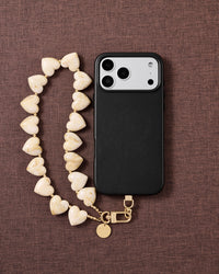 Ivory marbled heart bead strap looped beside a black phone case on brown fabric, gold hook and engraved disc resting at the base.
