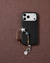 Flat-lay on brown fabric: a black phone with the silver-tone star, gold-striped blue bead and tortoiseshell cube strap on tan cord.