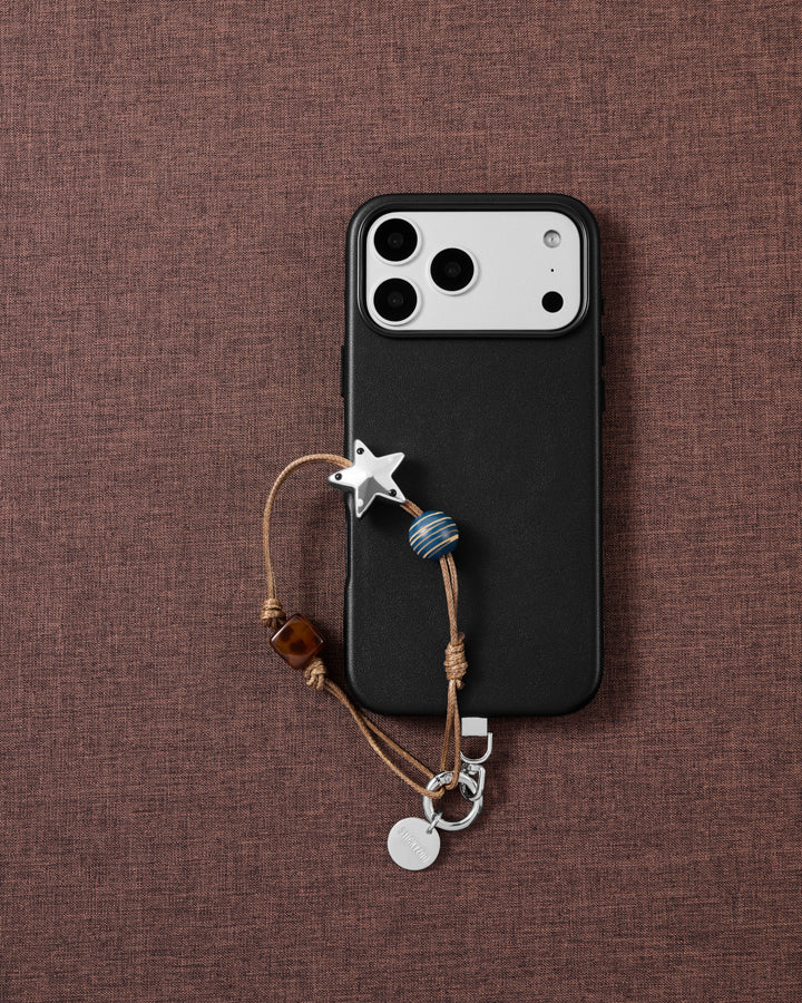 Flat-lay on brown fabric: a black phone with the silver-tone star, gold-striped blue bead and tortoiseshell cube strap on tan cord.