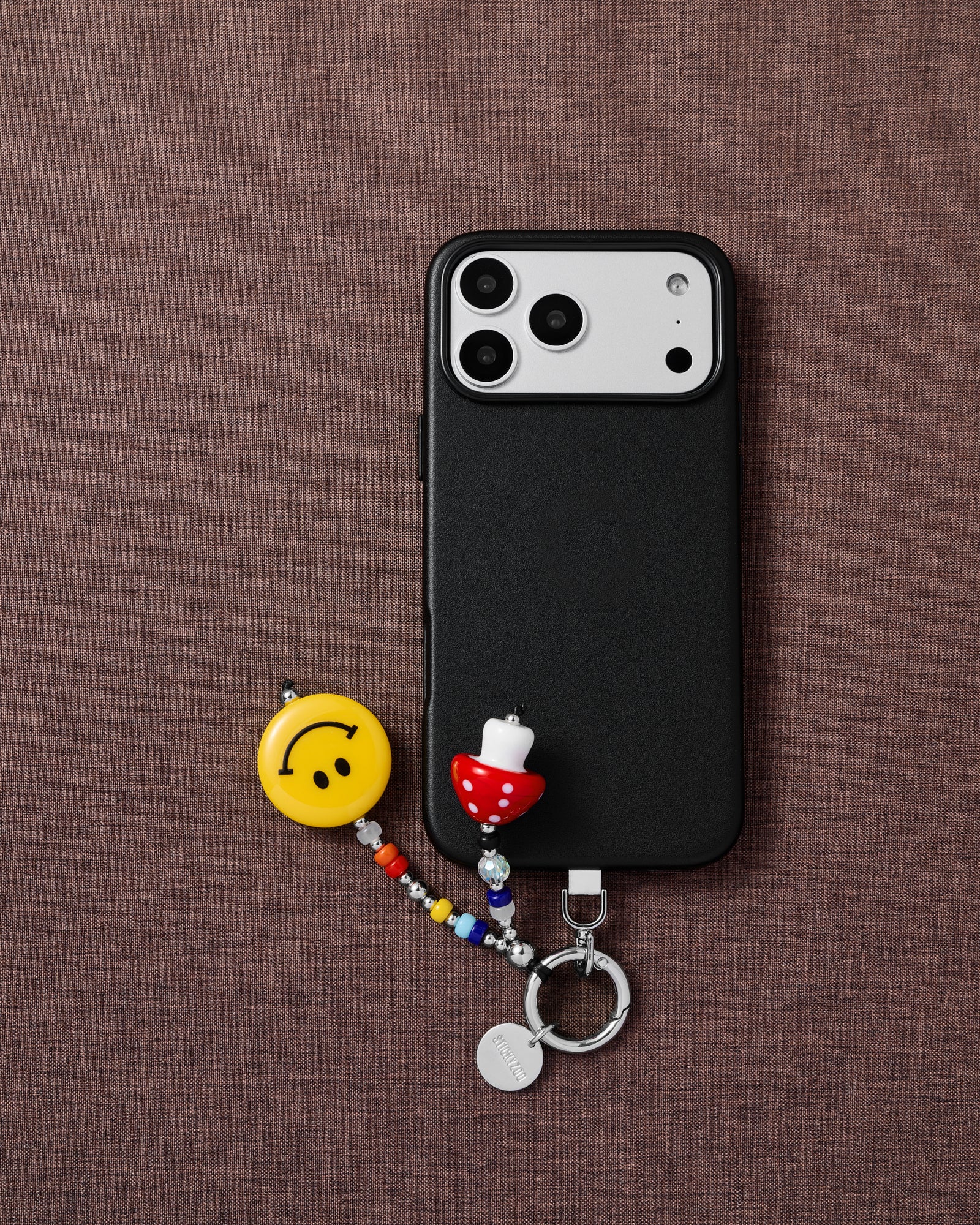 {{Designed in Amsterdam}} Phone with smiley mushroom charm on brown textile flat-lay showing STICKYZOO phone charm styling