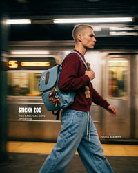 Male model walking past subway wearing STICKYZOO Capster Backpack, dalmatian-spotted faux suede with charm bundle