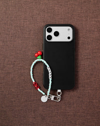Cherry RRR beaded phone strap with black phone on dark brown fabric lifestyle flat-lay