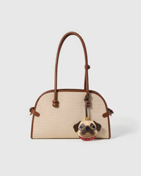 The Pug Charm styled on a cream canvas-and-tan top-handle bag, front — the exclusive piece that changes the whole energy of a bag.
