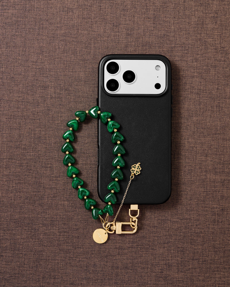 DESIGNED IN AMSTERDAM — Emerald clover heart phone strap styled flat-lay with gold four-leaf clover charm and emerald beads — Rated 4.9 stars