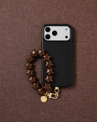 Designed in Amsterdam — cocoa star phone strap and black phone case on a deep cocoa fabric lifestyle flat-lay