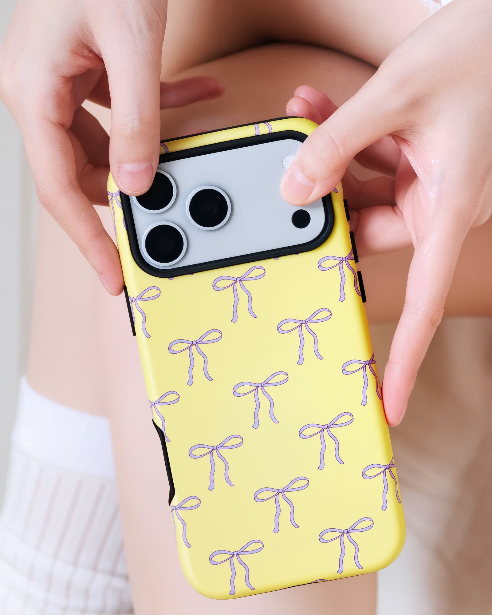 Two hands holding the butter-yellow bow phone case during the day with the lilac bows clearly visible