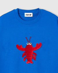 Close-up of plush shrimp applique showing red body, googly eyes, claws, and antennae on blue cotton