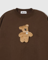 Close-up of plush teddy bear with pearl necklace and ear bow on brown cotton sweatshirt showing 3D texture