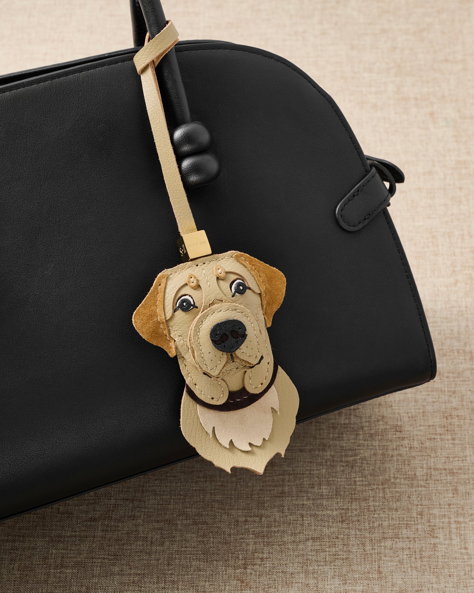 {{A detail people notice.}} Labrador Retriever charm close on a glossy black top-handle bag — the warm face that makes strangers stop and say it looks exactly like their dog.