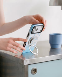 Phone standing hands-free on a counter on the ring kickstand of the STICKYZOO sky-star case beside a blue mug