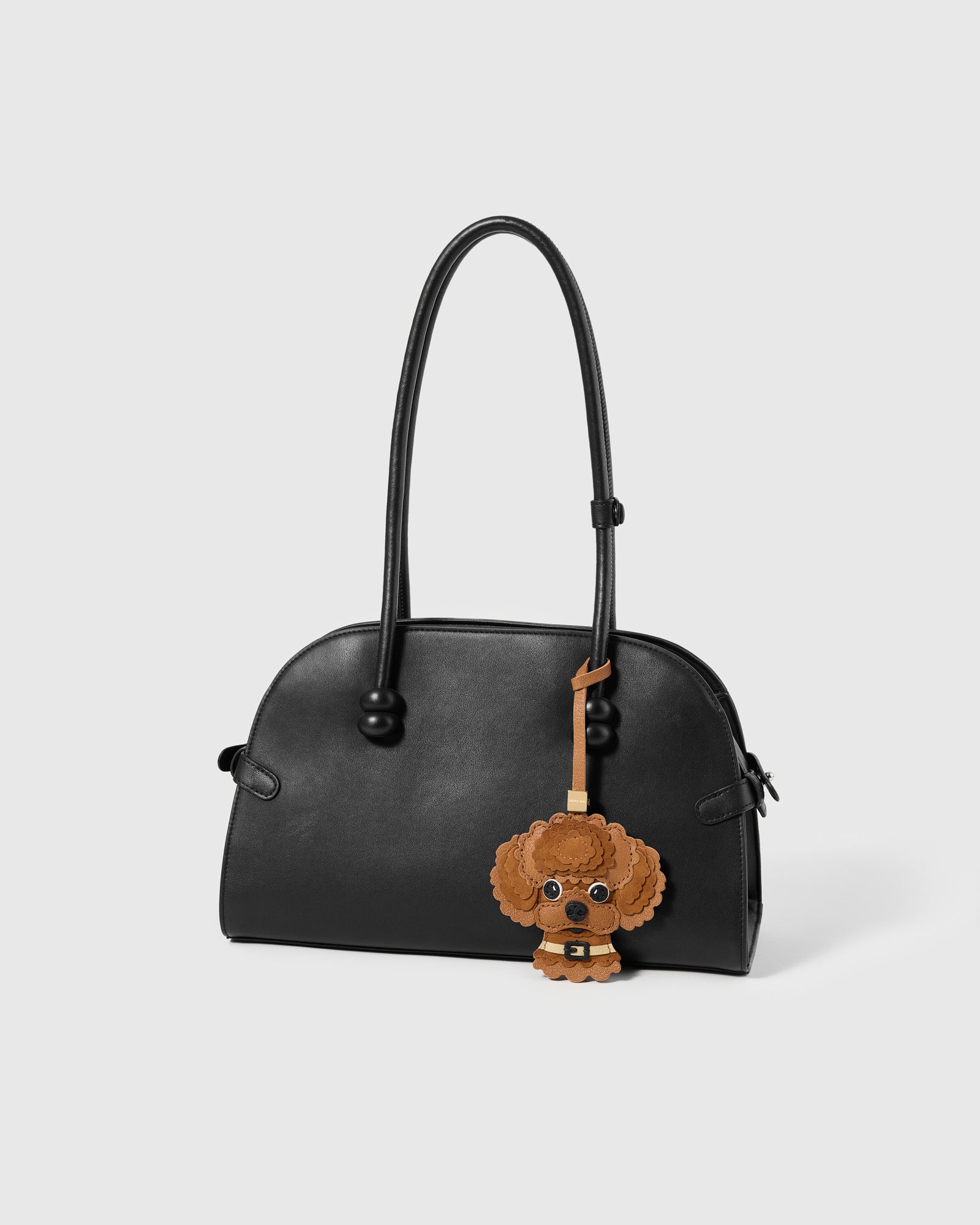 {{"People stop me to ask about it." — Mia K.}} The Poodle Charm on a glossy black bag, side angle — French Alran goatskin × Italian Alcantara. 7 days per piece.