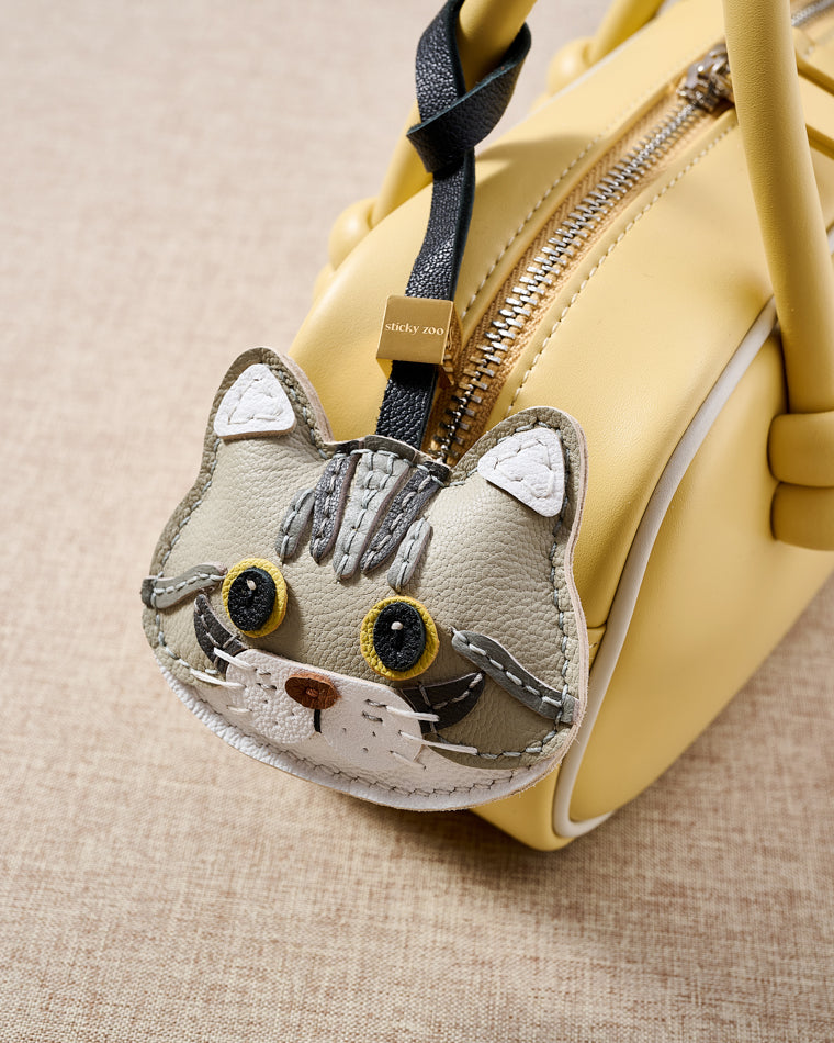 {{5,000+ Reviews.}} Silver Tabby American Shorthair hanging on a soft-yellow box bag, close detail — 5,000+ reviews across STICKYZOO. Real French Alran leather, 3 days by hand.